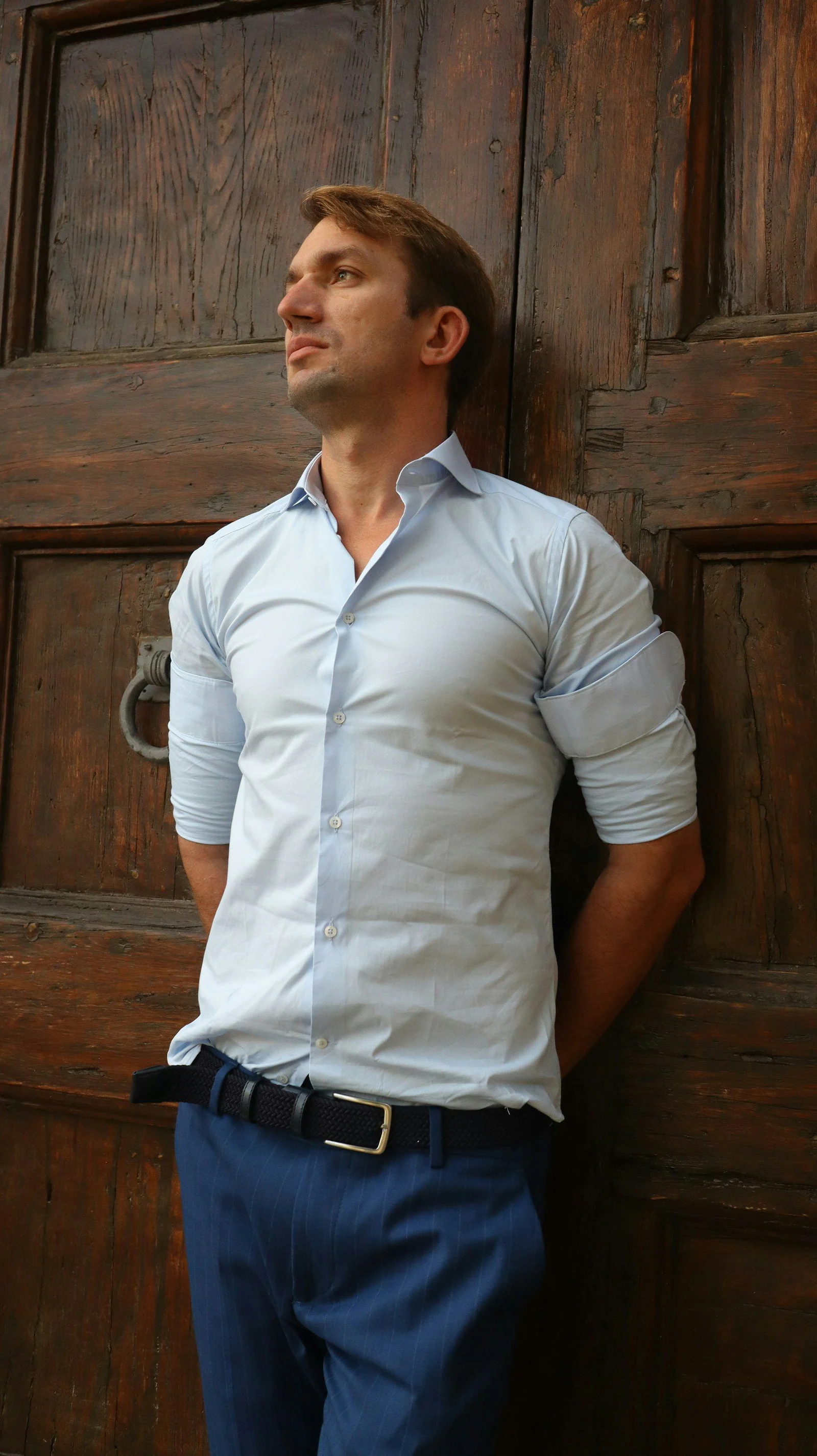 A man leaning against a wooden door wearing a white shirt and blue pants